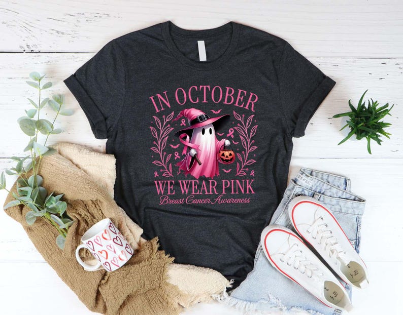 May include: Dark gray t-shirt with the text "IN OCTOBER WE WEAR PINK Breast Cancer Awareness". The shirt features a pink ghost wearing a witch hat and holding a pumpkin. A coffee mug with heart designs and white sneakers are also visible.