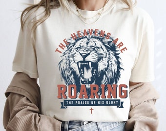 May include: Cream-colored t-shirt featuring a roaring lion graphic in navy blue and red. The text reads "THE HEAVENS ARE ROARING" above and "THE PRAISE OF HIS GLORY" below the lion, with a cross symbol.