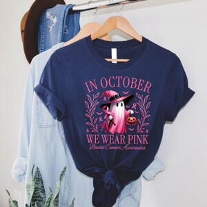 May include: Navy blue t-shirt with the text "IN OCTOBER WE WEAR PINK Breast Cancer Awareness". The shirt features a ghost wearing a pink witch hat and holding a pink ribbon and a jack-o'-lantern. The design is surrounded by pink leaves and ribbons.