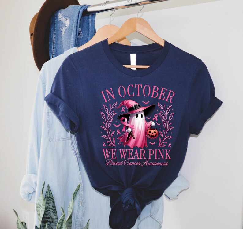May include: Navy blue t-shirt with the text "IN OCTOBER WE WEAR PINK Breast Cancer Awareness". The shirt features a ghost wearing a pink witch hat and holding a pink ribbon and a jack-o'-lantern. The design is surrounded by pink leaves and ribbons.
