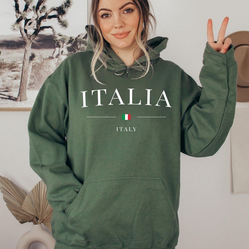 May include: A green hoodie with the word "ITALIA" in white block letters. Below the word is a horizontal line with the Italian flag in the center. The word "ITALY" is printed below the flag. The hoodie has a front pocket and a drawstring hood.
