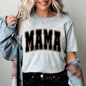 Może przedstawiać: Szary T-shirt z czarnym nadrukiem "MAMA". Litery są obwiedzione wzorem w panterkę.