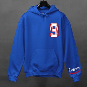 K&ouml;nnte beinhalten: Royalblauer Kapuzenpullover mit einer wei&szlig;en Baseballnummer 9 mit roten N&auml;hten. Das Wort "Custom" ist in Rot mit roten N&auml;hten auf dem &Auml;rmel gestickt.