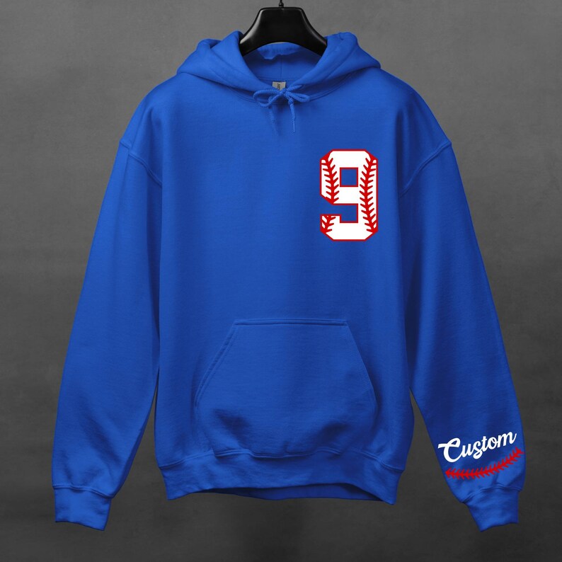 K&ouml;nnte beinhalten: Royalblauer Kapuzenpullover mit einer wei&szlig;en Baseballnummer 9 mit roten N&auml;hten. Das Wort "Custom" ist in Rot mit roten N&auml;hten auf dem &Auml;rmel gestickt.