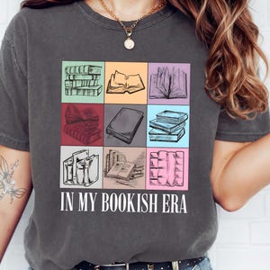 K&ouml;nnte beinhalten: Dunkelgraues T-Shirt mit einem farbenfrohen Muster von skizzierten B&uuml;chern in einem Raster. Der Text "IN MY BOOKISH ERA" ist darunter in Wei&szlig; gedruckt. Das Shirt ist ein bequemer, l&auml;ssiger Stil.