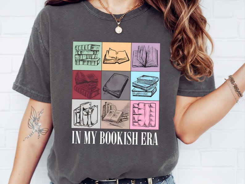 K&ouml;nnte beinhalten: Dunkelgraues T-Shirt mit einem farbenfrohen Muster von skizzierten B&uuml;chern in einem Raster. Der Text "IN MY BOOKISH ERA" ist darunter in Wei&szlig; gedruckt. Das Shirt ist ein bequemer, l&auml;ssiger Stil.