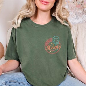 May include: A green t-shirt with a retro-style graphic that says "Spelling Bee Mom's Club" with a cartoon ghost wearing a crown.