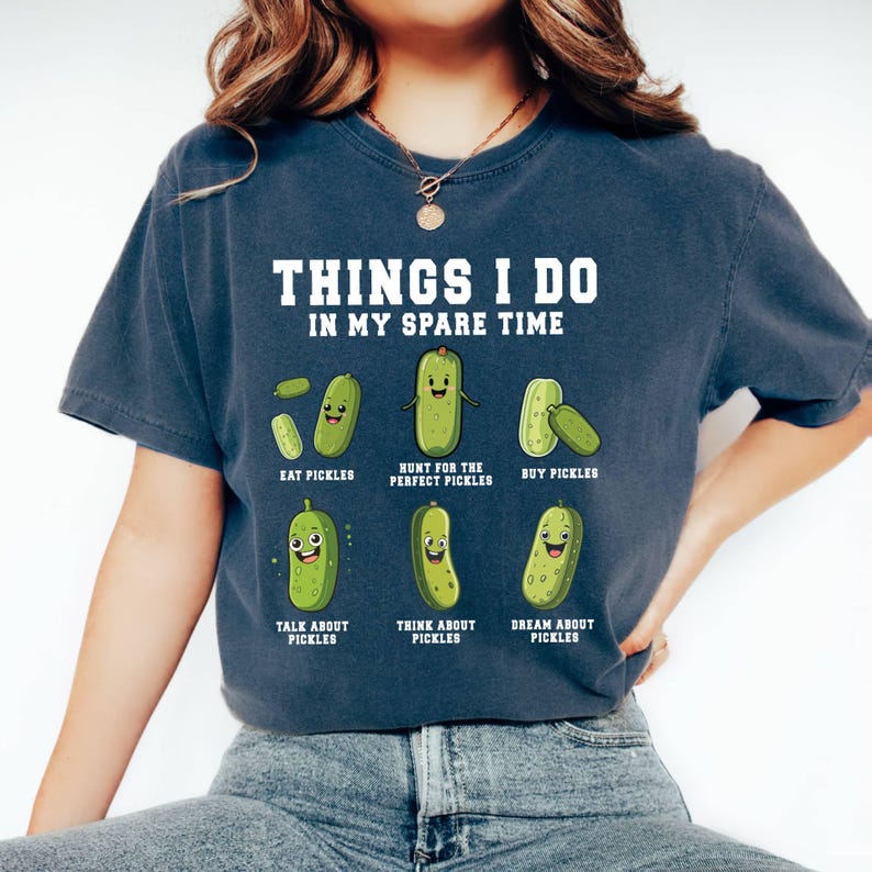 Pu&ograve; includere: Una maglietta blu navy con una grafica bianca che dice "Things I do in my spare time" e presenta sei cetrioli di cartone animato con espressioni diverse. Il testo sotto ogni cetriolo descrive attivit&agrave; legate ai cetrioli, come "Mangiare cetrioli", "Cacciare i cetrioli perfetti", "Comprare cetrioli", "Parlare di cetrioli", "Pensare ai cetrioli" e "Sognare i cetrioli".