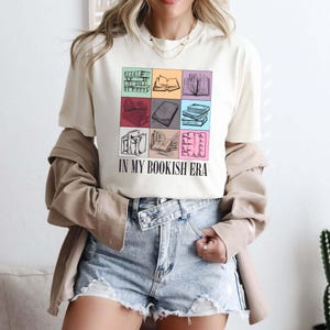 K&ouml;nnte beinhalten: Cremefarbenes T-Shirt mit einem Raster aus illustrierten B&uuml;chern in verschiedenen Farben. Der Text "IN MY BOOKISH ERA" ist darunter gedruckt. Die Person tr&auml;gt Jeansshorts und eine beige Jacke.