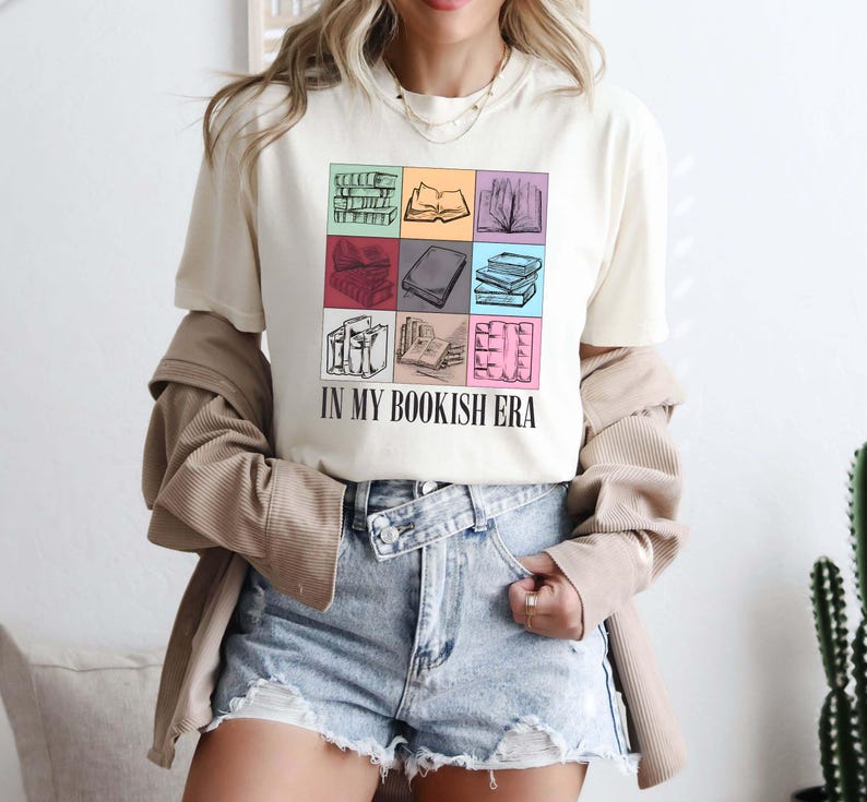 K&ouml;nnte beinhalten: Cremefarbenes T-Shirt mit einem Raster aus illustrierten B&uuml;chern in verschiedenen Farben. Der Text "IN MY BOOKISH ERA" ist darunter gedruckt. Die Person tr&auml;gt Jeansshorts und eine beige Jacke.