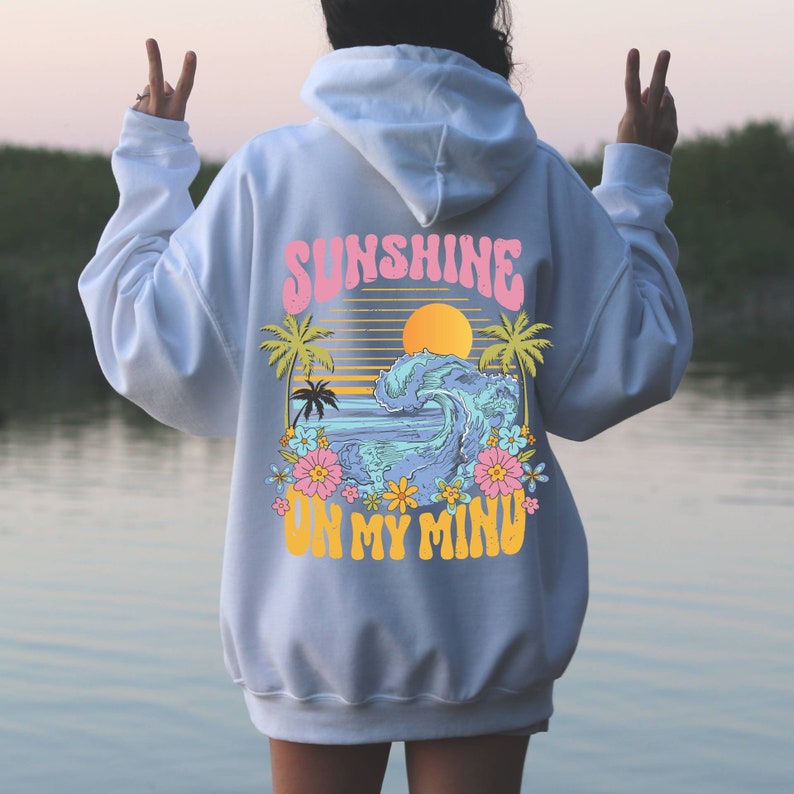 Puede incluir: Sudadera con capucha blanca con un gr&aacute;fico colorido en la espalda. El gr&aacute;fico presenta las palabras "SUNSHINE ON MY MIND" en una fuente retro, junto con una escena de playa con una ola, palmeras y flores.