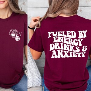 May include: Burgundy t-shirt with a white graphic on the front of a can with a skeleton hand and the text "++" and a white graphic on the back that says "Fueled by Energy Drinks & Anxiety".