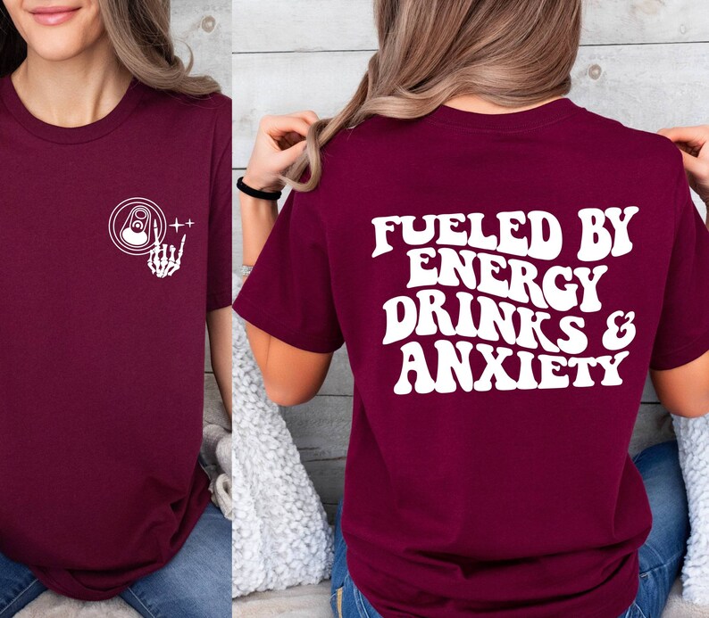 May include: Burgundy t-shirt with a white graphic on the front of a can with a skeleton hand and the text "++" and a white graphic on the back that says "Fueled by Energy Drinks & Anxiety".