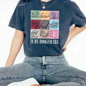 K&ouml;nnte beinhalten: Marineblaues T-Shirt mit einem farbenfrohen Raster von Buchillustrationen. Der Text "IN MY BOOKISH ERA" ist in Wei&szlig; gedruckt. Das Design umfasst B&uuml;cherstapel, offene B&uuml;cher und Buchr&uuml;cken in verschiedenen Farben.