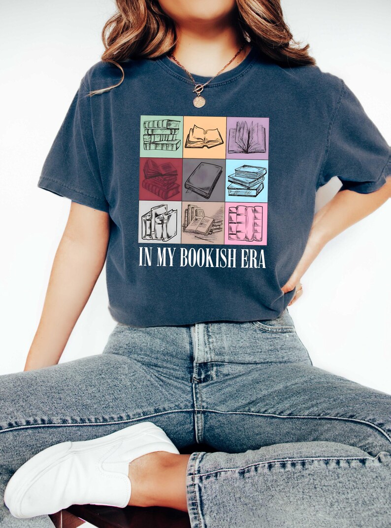 K&ouml;nnte beinhalten: Marineblaues T-Shirt mit einem farbenfrohen Raster von Buchillustrationen. Der Text "IN MY BOOKISH ERA" ist in Wei&szlig; gedruckt. Das Design umfasst B&uuml;cherstapel, offene B&uuml;cher und Buchr&uuml;cken in verschiedenen Farben.