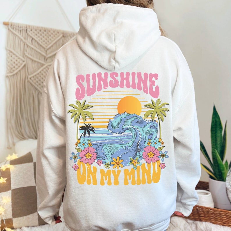 May include: A white hoodie with a graphic print of a sunset, palm trees, a wave, and flowers. The text 'Sunshine on my mind' is printed in pink and yellow letters.