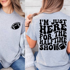 May include: Gray t-shirt with a football graphic on the left side and the text "I'm Just Here For The Halftime Show" with a football graphic on the right side.