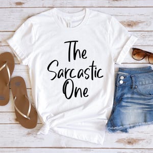 Peut inclure: T-shirt blanc avec un texte noir qui dit "The Sarcastic One". Le t-shirt est pos&eacute; sur une surface en bois avec une paire de sandales marrons et un short en jean bleu.
