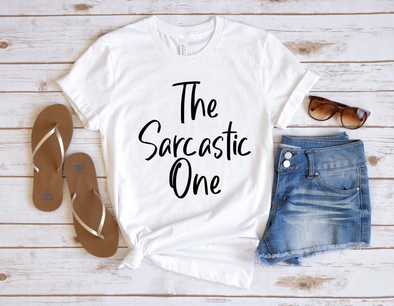 Peut inclure: T-shirt blanc avec un texte noir qui dit "The Sarcastic One". Le t-shirt est pos&eacute; sur une surface en bois avec une paire de sandales marrons et un short en jean bleu.