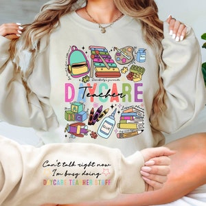 May include: A beige sweatshirt with a colorful graphic design of daycare items, including a backpack, xylophone, paint palette, crayons, glue, books, and a toy airplane. The text "Somebody's favorite DAYCARE Teacher" is printed on the sweatshirt. The bottom of the sweatshirt has the text "Can't talk right now I'm busy doing DAYCARE TEACHER STUFF."