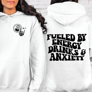 Puede incluir: Sudadera con capucha blanca con un gr&aacute;fico negro en la parte delantera de una lata con una mano de esqueleto haciendo el signo de rock and roll. La parte posterior de la sudadera tiene el texto "Fueled by Energy Drinks & Anxiety" en una fuente retro.