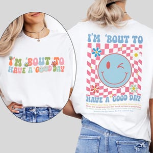 May include: White t-shirt with the text "I'M 'BOUT TO HAVE A GOOD DAY" in colorful, retro-style lettering. The back features a smiley face, flowers, and a checkered pattern. The shirt has a relaxed fit.
