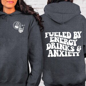 Puede incluir: Una sudadera gris oscura con un gr&aacute;fico blanco en la parte delantera de una mano de esqueleto sosteniendo una lata de bebida energ&eacute;tica. La parte posterior de la sudadera tiene el texto "Fueled by Energy Drinks & Anxiety" en blanco.