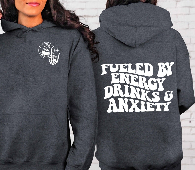 Puede incluir: Una sudadera gris oscura con un gr&aacute;fico blanco en la parte delantera de una mano de esqueleto sosteniendo una lata de bebida energ&eacute;tica. La parte posterior de la sudadera tiene el texto "Fueled by Energy Drinks & Anxiety" en blanco.