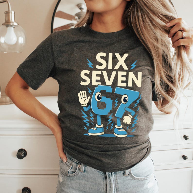 May include: Dark gray t-shirt with the words "SIX SEVEN" above a cartoon-style "67" in light blue. The "6" has a hand, and both numbers have lightning bolt accents. The shirt is a casual style.