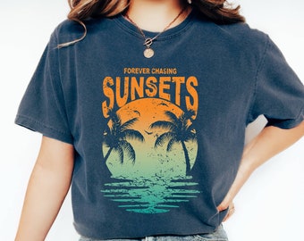May include: A navy blue t-shirt with the words "FOREVER CHASING SUNSETS" in orange. The graphic features a sunset scene with palm trees and water. The shirt is a relaxed fit.