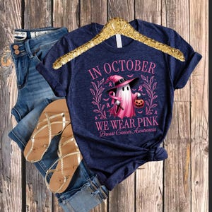 May include: Navy blue t-shirt with the text "IN OCTOBER WE WEAR PINK Breast Cancer Awareness" and a pink ghost graphic holding a jack-o'-lantern. The shirt is displayed with blue denim shorts and gold sandals.