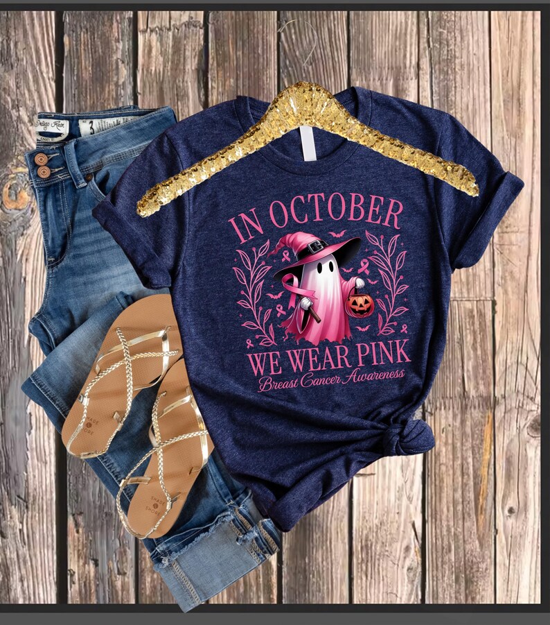 May include: Navy blue t-shirt with the text "IN OCTOBER WE WEAR PINK Breast Cancer Awareness" and a pink ghost graphic holding a jack-o'-lantern. The shirt is displayed with blue denim shorts and gold sandals.