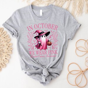 May include: A light gray t-shirt with a pink graphic for Breast Cancer Awareness. The graphic features a ghost wearing a witch hat and holding a jack-o'-lantern. The text reads "In October We Wear Pink".
