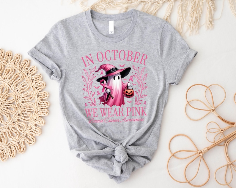 May include: A light gray t-shirt with a pink graphic for Breast Cancer Awareness. The graphic features a ghost wearing a witch hat and holding a jack-o'-lantern. The text reads "In October We Wear Pink".