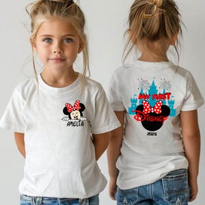 May include: White t-shirt featuring a Minnie Mouse graphic with a red polka dot bow and the name "AMELIA" in black. The back of the shirt has the text "MY FIRST DISNEY" with a castle and fireworks design, and the year "2025".