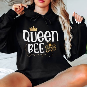 May include: Black hooded sweatshirt with the text "Queen Bee" in white and gold. The design includes a gold crown and a bee illustration. The sweatshirt is a casual garment.