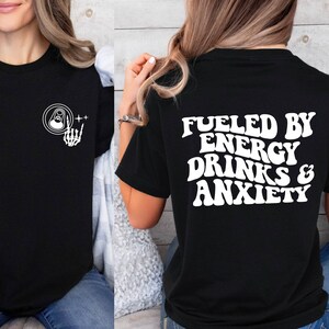 May include: Black t-shirt with white text that reads "Fueled by Energy Drinks & Anxiety". The front of the shirt has a white graphic of a skeleton hand holding a can.