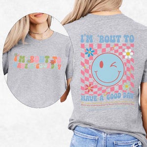 May include: A light gray t-shirt with the text "I'M BOUT TO HAVE A GOOD DAY" in colorful, retro-style lettering. The back of the shirt features a smiley face and a checkered pattern.