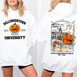 May include: White hooded sweatshirt with a Halloween theme. The front features a pumpkin graphic with the text "Halloweentown University Est. 1998." The back has a detailed illustration of a pumpkin, building, and the text "Halloweentown Est. 1998."