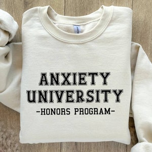 May include: A beige sweatshirt with black text that reads "Anxiety University - Honors Program - "