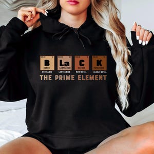 Puede incluir: Sudadera con capucha negra con un estampado gr&aacute;fico de la tabla peri&oacute;dica de los elementos para las letras "B", "La", "C" y "K" que representan la palabra "Black". El texto "The Prime Element" est&aacute; impreso debajo del gr&aacute;fico.