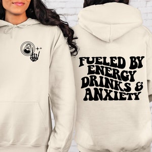 Puede incluir: Una sudadera con capucha de color crema con un gr&aacute;fico negro de una lata y una mano de esqueleto en la parte delantera y el texto "Fueled by Energy Drinks & Anxiety" en la parte posterior.