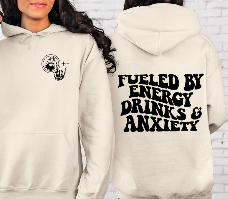 Puede incluir: Una sudadera con capucha de color crema con un gr&aacute;fico negro de una lata y una mano de esqueleto en la parte delantera y el texto "Fueled by Energy Drinks & Anxiety" en la parte posterior.