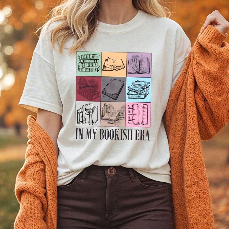 K&ouml;nnte beinhalten: Cremefarbenes T-Shirt mit einem bunten Raster von Buchillustrationen. Der Schriftzug "IN MY BOOKISH ERA" ist darunter gedruckt. Die Person tr&auml;gt eine rostorangefarbene Strickjacke.