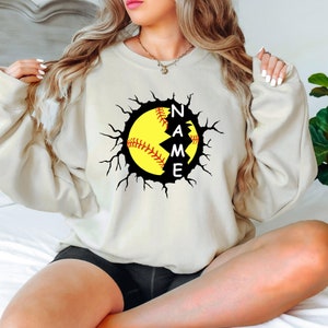 May include: A cream-colored sweatshirt with a graphic of a softball breaking through a cracked black circle. The word "NAME" is written vertically inside the circle.