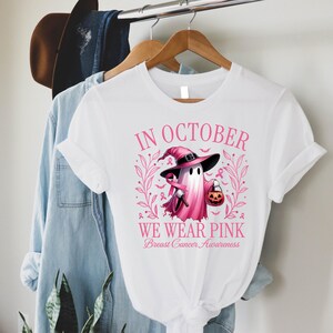 May include: White t-shirt with a pink graphic for Breast Cancer Awareness. The graphic features a ghost wearing a witch hat and holding a pumpkin. The text reads "In October We Wear Pink".