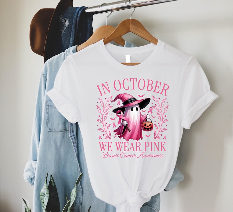 May include: White t-shirt with a pink graphic for Breast Cancer Awareness. The graphic features a ghost wearing a witch hat and holding a pumpkin. The text reads "In October We Wear Pink".