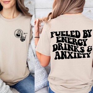 May include: A beige t-shirt with a black graphic of a skeleton hand holding a can of energy drink on the front and the text "Fueled by Energy Drinks & Anxiety" on the back.
