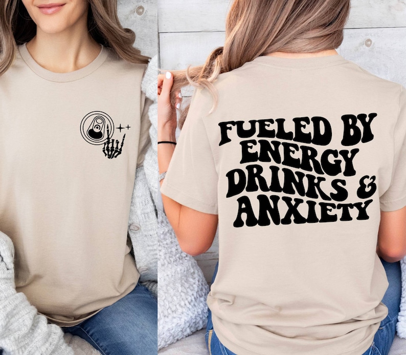May include: A beige t-shirt with a black graphic of a skeleton hand holding a can of energy drink on the front and the text "Fueled by Energy Drinks & Anxiety" on the back.