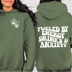 Puede incluir: Una sudadera con capucha verde con el texto "Fueled by Energy Drinks & Anxiety" en blanco en la espalda. La parte delantera de la sudadera tiene un gr&aacute;fico blanco de una lata con una mano de esqueleto haciendo el signo de rock and roll.